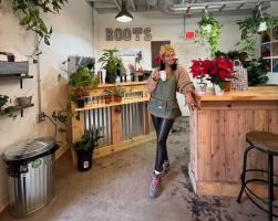 Owner, Bee, leans on a counter in a room filled with flowers at her business Roots Plants & Coffee in Olde Towne Slidell